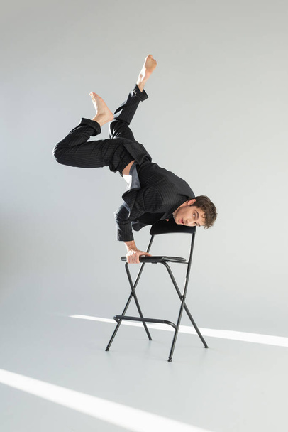 full length of stylish barefoot man in black suit looking at camera while doing trick on chair on grey background - Photo, image
