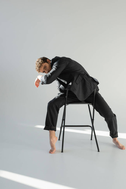 full length of barefoot man in black and striped suit posing on chair and looking at camera on grey background - Фото, изображение