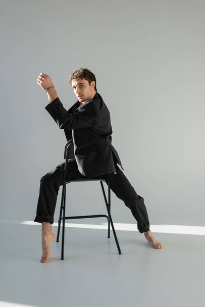 full length of barefoot man in black stylish blazer and silver bracelet posing on chair and looking at camera on grey background - Foto, immagini