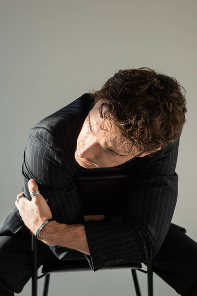 брюнетка мужчина в черной полосатой блейзер и металлический браслет позирует сидя на стуле со скрещенными руками изолированы на сером - Фото, изображение