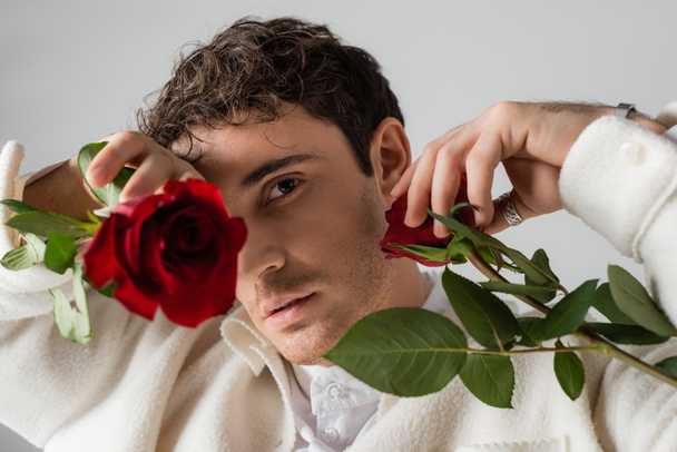 stylish man in white clothes obscuring face with red roses while looking at camera isolated on grey - Foto, Bild