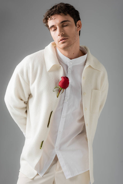 trendy man in white jacket decorated with red roses standing with closed eyes isolated on grey - Foto, imagen