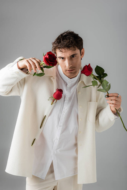 man wearing white stylish clothes looking at camera while posing with red roses isolated on grey - Fotó, kép