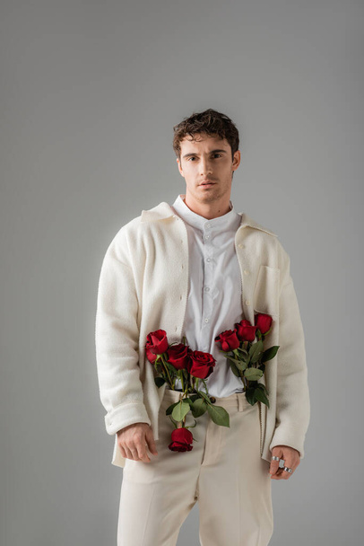 front view of man in white stylish clothes standing with red roses in pants isolated on grey - Fotó, kép