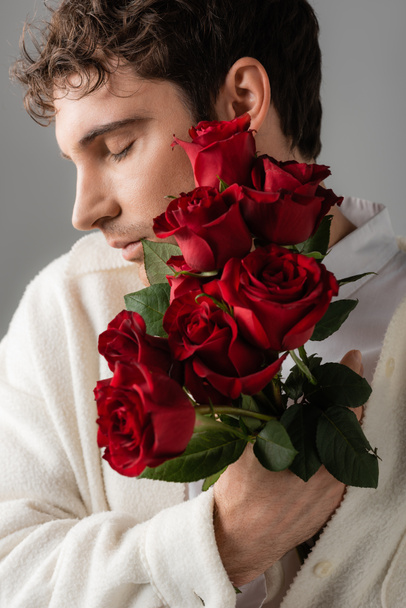 мужчина с закрытыми глазами держит букет из красных свежих роз, изолированных на сером - Фото, изображение