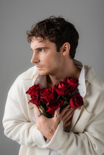 брюнетка мужчина в белой мягкой куртке и серебряные кольца пальца держа букет красных роз и глядя в сторону изолированы на сером - Фото, изображение