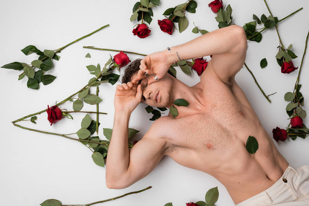 вид сверху на мускулистого мужчину без рубашки с закрытыми глазами, лежащего рядом с красными розами на белом фоне - Фото, изображение