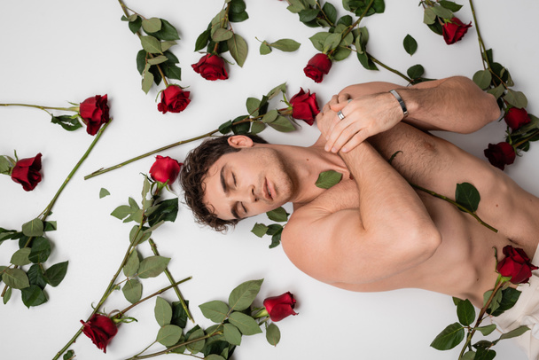 top view of shirtless man in silver bracelet and finger ring lying with closed eyes near red roses on white background - Fotografie, Obrázek