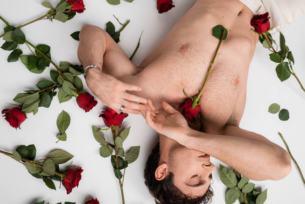 top view of sexy man with shirtless torso and closed eyes lying near red roses on white background - Photo, image
