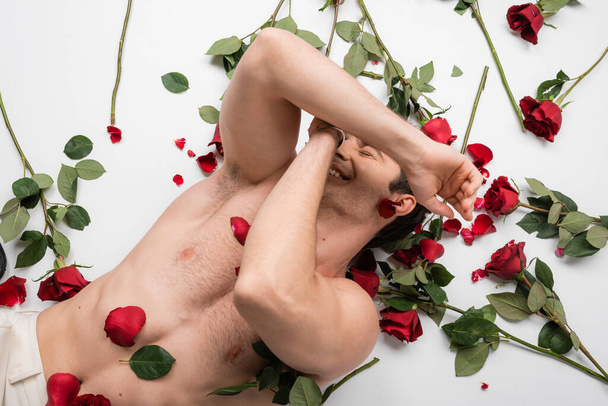 κορυφαία άποψη του γυμνού άνδρα με μυϊκό κορμό σκιάζει το πρόσωπο και χαμογελά κοντά κόκκινα τριαντάφυλλα σε λευκό φόντο - Φωτογραφία, εικόνα