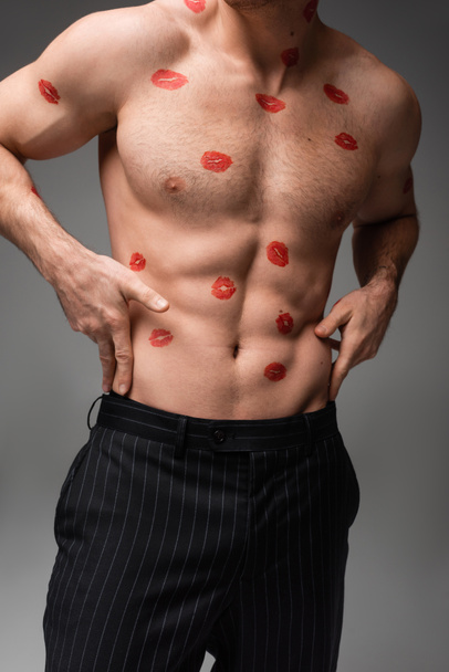 cropped view of man in black pants touching muscular torso with red lip prints isolated on grey - Photo, Image