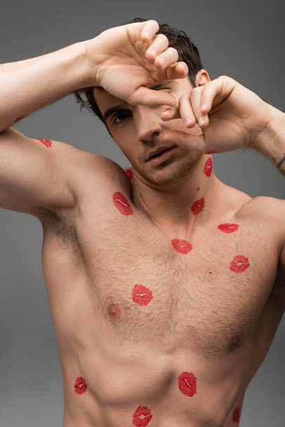 sexy shirtless man with red lipstick prints on torso obscuring face with hands while looking at camera isolated on grey - Foto, afbeelding
