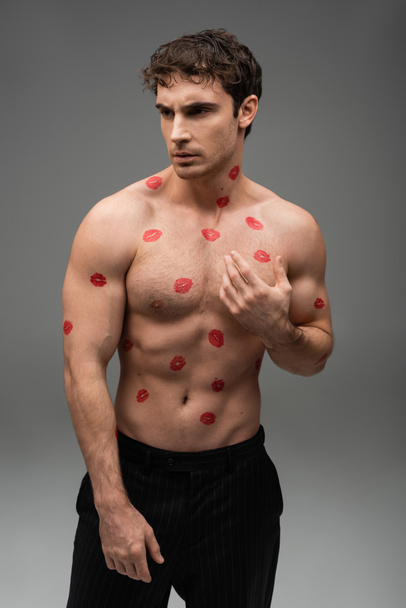 brunette muscular man with red lipstick prints on shirtless body looking away on grey background - Photo, image