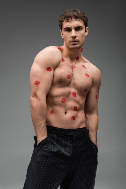 sexy athletic man with red kiss prints on body standing with hands in pockets of black trousers on grey background - Foto, Imagen