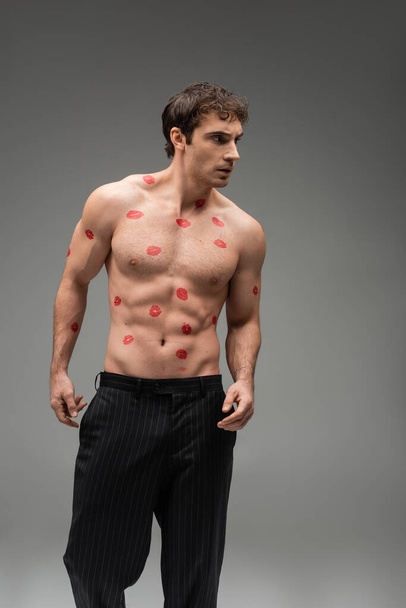 sexy man in black pants posing with red kiss prints on shirtless muscular torso and looking away on grey background - Фото, изображение