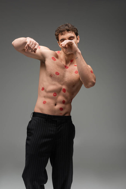 athletic man with red kisses on shirtless body obscuring face while posing with closed eyes on grey background - Photo, image