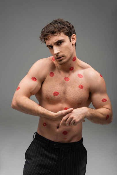 athletic man with red lip prints on shirtless muscular body looking at camera on grey background - Foto, afbeelding