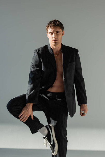 sexy brunette man in black pants and blazer on shirtless body standing on one leg and looking at camera on grey background - Photo, image