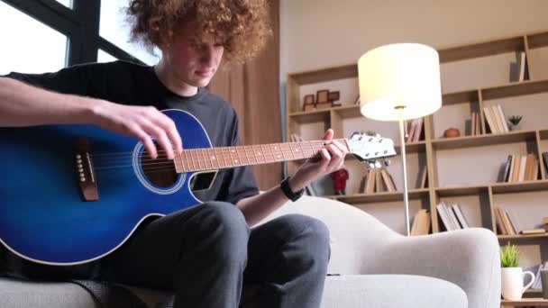 A curly-haired guy is sitting on a sofa and playing an acoustic guitar, he has a headache. Migraine and headache in a young man - Materiał filmowy, wideo