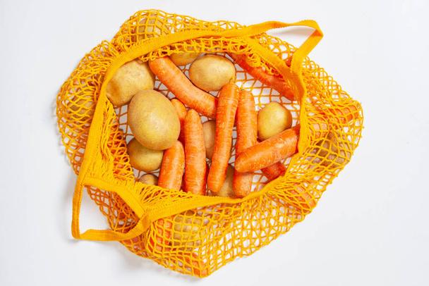Orange mesh bag. View from above. Close-up. Fresh vegetables from the farmers market. Raw potatoes, raw carrots. Conscious consumption, reusable bag, string bag. Vegetarianism, raw food diet, ecology, fresh vegetables. On a white background. open bag - Foto, Bild