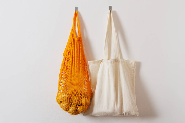 mesh grocery bag. Reusable bag. Vegetarianism, raw food diet, conscious consumption, grid. orange mesh bag hanging on a hook. Several shopping bags. White, yellow. Raw potatoes - Zdjęcie, obraz