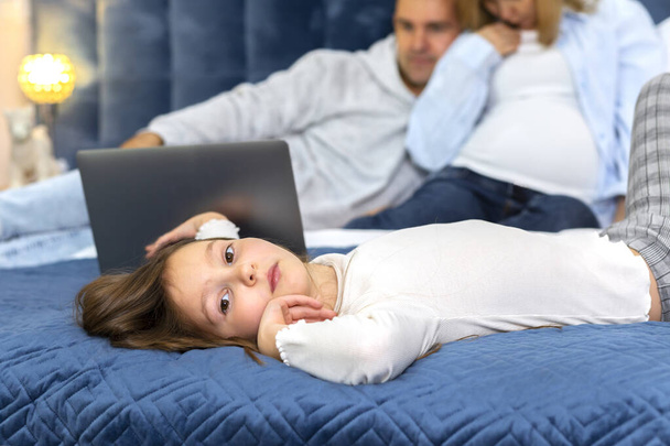Sad teen girl lays on bed on foreground and looks at camera while dad and pregnant mom are watching video on gadget on background. Raising a teen, feeling lonely. being jealous to sibling. Horizontal - Фото, изображение