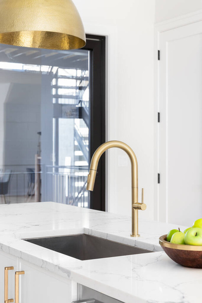A gold kitchen faucet in a white kitchen with a marble countertop, gold light fixture hanging down, and a bowl of green apples. - Zdjęcie, obraz
