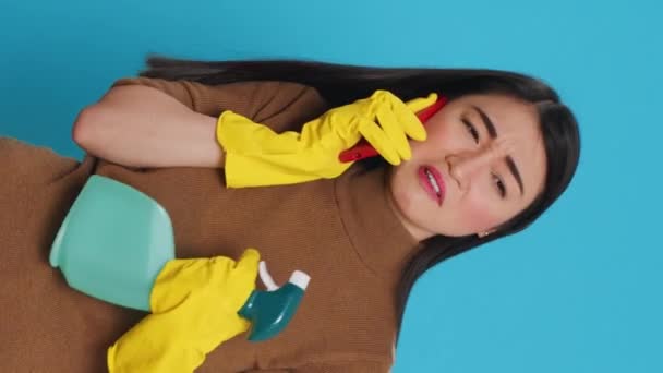 Вертикальное видео: азиатская домохозяйка в защитных перчатках, держа бутылку с моющим средством в руках, разговаривая на мобильном телефоне с дистанционным другом, стоящим в студии на синем фоне. Уборка и - Кадры, видео