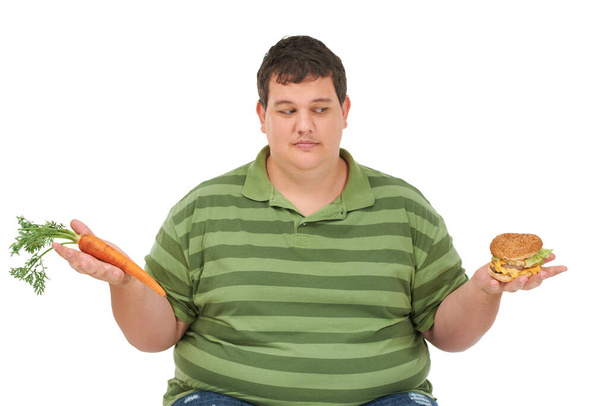 Заманчиво попробовать. Толстый молодой человек, сидящий с морковкой в одной руке, бургер в другой и смотрящий на бургер с заманчивым выражением лица - Фото, изображение