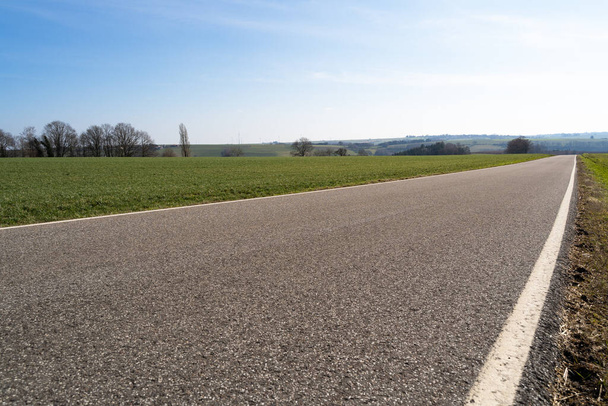 Low angle view of a paved rural road between grassfields in the landscape with blue sunny sky in spring - Foto, imagen