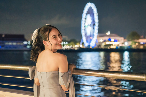 Happy beautiful young asian woman in dress smiling and sightseeing illuminated ferris wheel during cruise ship on the river in the night at Bangkok - Foto, imagen