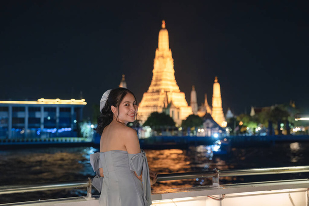 Прекрасна молода азійка в сукні стоїть і дивиться на фотоапарат на палубі, а Ват Арун світиться вночі. Їжа, що пливе по річці Чао Фрая (Бангкок). - Фото, зображення