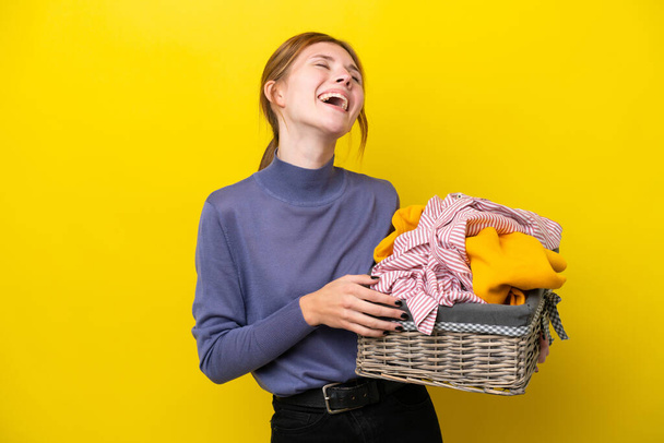 Νεαρή Αγγλίδα που κρατάει καλάθι με ρούχα απομονωμένη σε κίτρινο φόντο γελώντας - Φωτογραφία, εικόνα