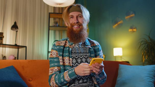 Hipster pelirroja barbudo hombre sentado en el sofá utiliza el teléfono móvil sonríe en la noche casa por la noche. Joven chico mensajes de texto compartir mensajes de contenido en aplicaciones de redes sociales de teléfonos inteligentes en línea viendo relajar película - Foto, imagen