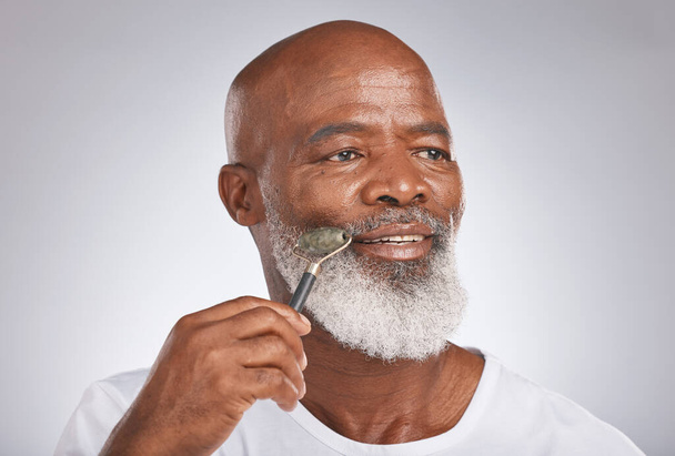 Senior black man, skincare and facial roller to massage skin during self care for wellness, dermatology and cosmetics. Headshot of male with beauty tool on grey studio background for a spa treatment. - Foto, imagen