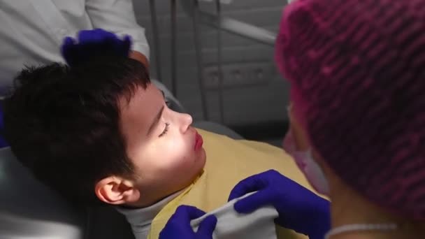 Lähikuva teini poika hammaslääkärit tuoli saada hampaiden hoito moderni pediatrinen hammashoitola. Taustakuva hammaslääkärin hygienististä pyyhkimässä pienen potilaan suuta hammastoimenpiteiden jälkeen. - Materiaali, video