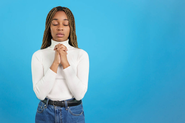 Studio portrait with blue background of an african woman with eyes closed and folded hands in prayer - Foto, afbeelding