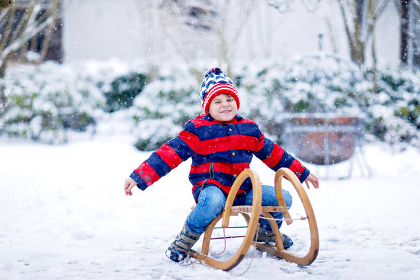 Маленький школьник развлекается катанием на санях во время снегопада. Счастливый ребенок катается на санках по снегу. Дошкольник катается на санях. Дети играют на свежем воздухе в снежном зимнем парке. Активное развлечение для семьи - Фото, изображение