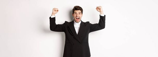 Επιτυχημένος όμορφος επιχειρηματίας θριαμβεύει, σηκώνει τα χέρια ψηλά και φωνάζει ναι, χαίρεται για την επίτευξη, στέκεται απέναντι σε λευκό φόντο. - Φωτογραφία, εικόνα
