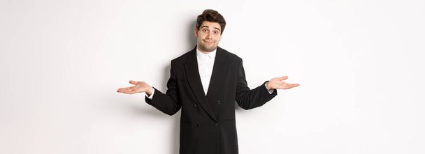 Image of handsome confused man in black suit, shrugging and looking clueless, dont know anything, standing over white background. - Photo, Image