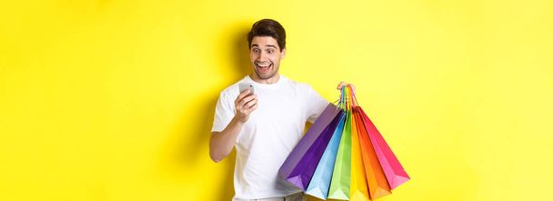 Alışveriş torbaları tutan heyecanlı bir adam cep telefonu ekranında mutlu görünüyor, sarı arka planda duruyor.. - Fotoğraf, Görsel