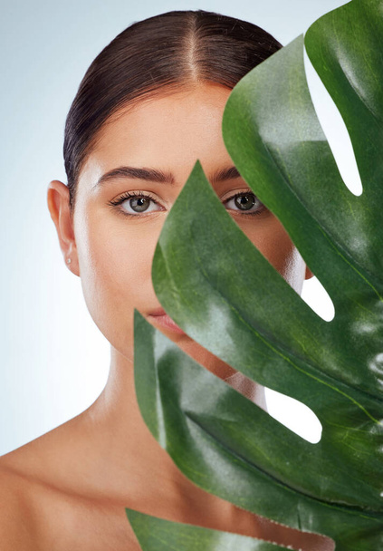 .Portrait of unknown woman covering her face with a green monstera plant leaf. Headshot of caucasian model posing against a grey background in a studio with smooth skin and a fresh healthy skincare. - Photo, Image