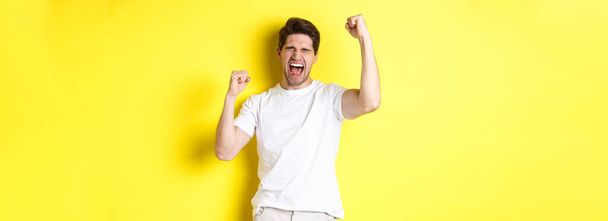 Excited handsome man winning and shouting for joy, raising hand up and triumping, achieve goal, standing over yellow background. - Photo, Image