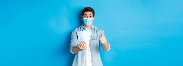 Concept de coronavirus, quarantaine et distanciation sociale. Jeune homme montrant des masques médicaux et pouce levé, debout sur fond bleu. - Photo, image