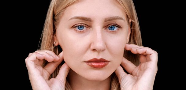 Plastic surgery, facelift, rejuvenation. Portrait of a beautiful middle aged woman looking at the camera, touching her cheekbones with her hands. Blue-eyed blonde caucasian girl on a dark background - Foto, imagen