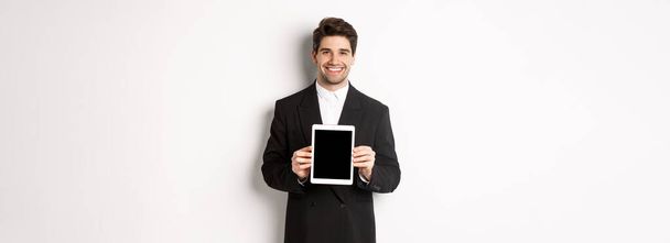 Portrait d'un bel homme d'affaires en costume tendance, montrant écran de tablette numérique et souriant, debout sur fond blanc. - Photo, image