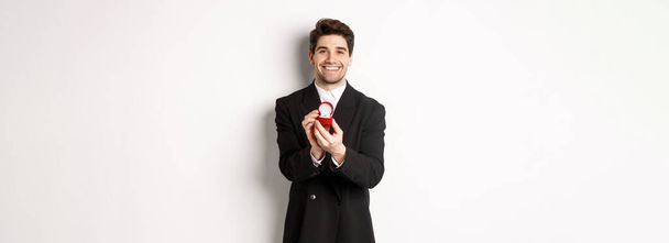 Obraz przystojnego chłopaka w czarnym garniturze składającego propozycję, proszącego o rękę i pokazującego obrączkę, stojącego nad białym tłem. - Zdjęcie, obraz