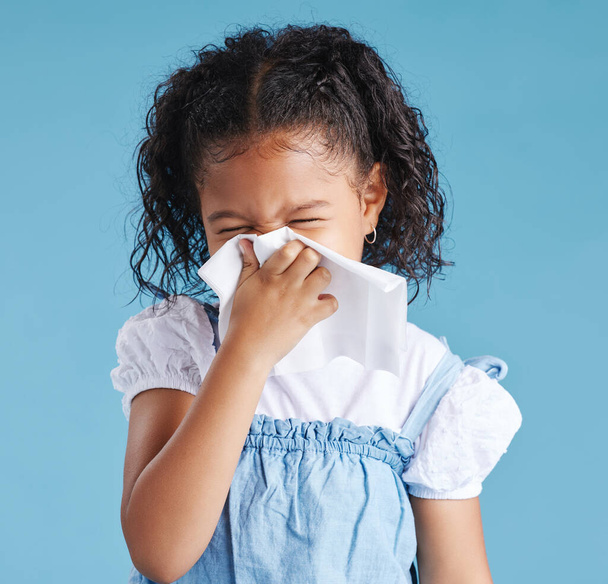 Little hispanic girl blowing her nose with tissue against blue studio background. Child feeling sick with flu virus or infection and suffering with snotty nose. - Fotoğraf, Görsel