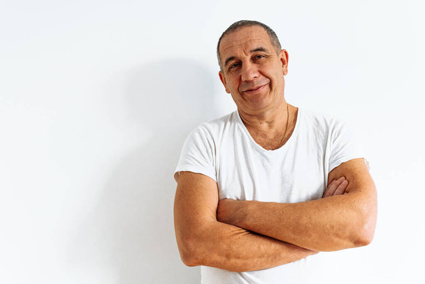 middle-aged man in white T-shirt with smile and positive emotion, facial wrinkles, smirk, against white wall. Emotional portrait of an adult man, facial expression, sincere joy - Photo, Image