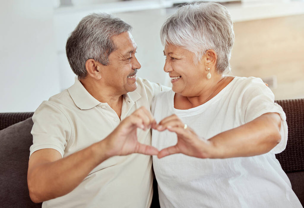 Χαμογελώντας μικτή φυλή ηλικιωμένο ζευγάρι κάνει σχήμα καρδιάς, σημάδι, σύμβολο με χειρονομία χέρι στο σπίτι. Ευτυχισμένοι Ισπανοί σύζυγοι που κάθονται μαζί στον καναπέ του σαλονιού. Ηλικιωμένοι συνταξιούχοι άνδρες και γυναίκες bondin. - Φωτογραφία, εικόνα